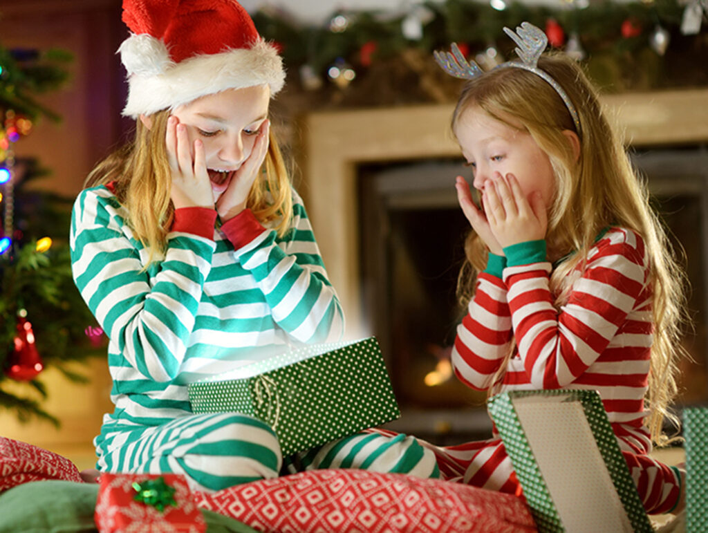 Happy little sisters wearing Christmas pajamas opening gift boxes by a fireplace in a cozy dark living room on Christmas eve.