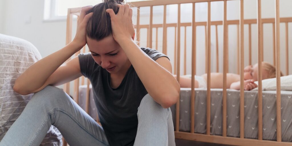 Mother holding head in hands suffering postnatal depression sitting next to baby sleeping in bed